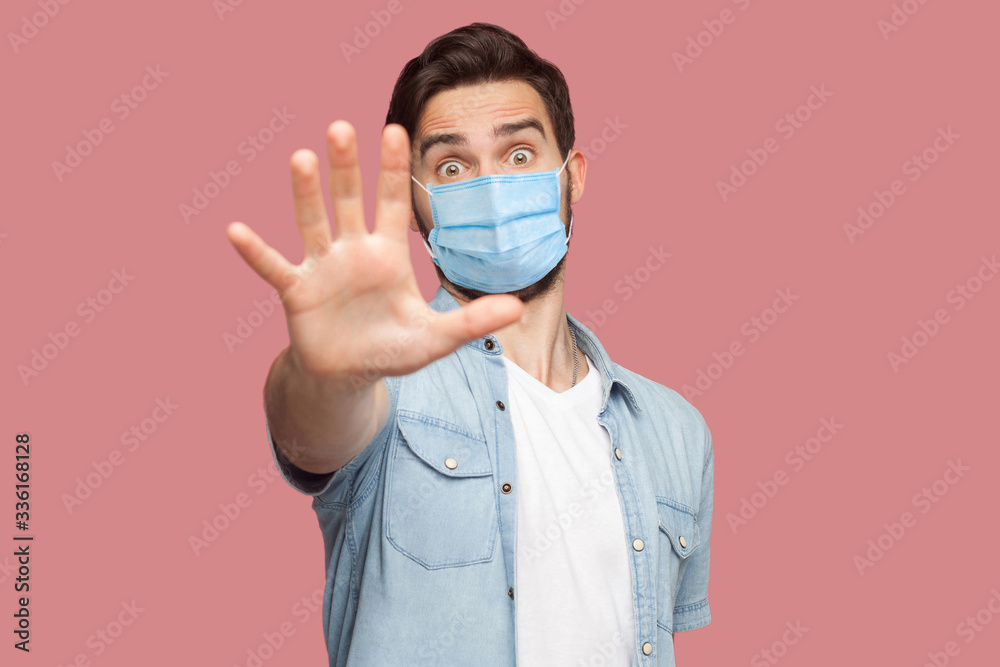 Stop, don't come any closer. . Portrait of angry young man with surgical medical mask in blue shirt standing with stop hand sign and looking at camera. indoor studio shot, isolated on pink background. Stop, don't come any closer. . Portrait of angry young man with surgical medical mask in blue shirt standing with stop hand sign and looking at camera. indoor studio shot, isolated on pink background.