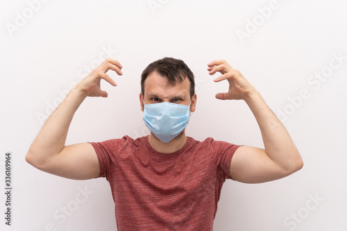 Caucasian man in facial surgical medical protective mask shows aggressive emotions with hands and angry eyes.