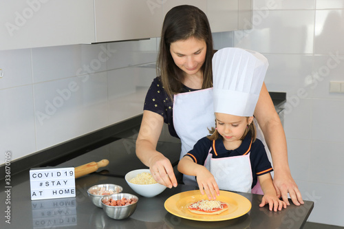 Young mother and her preschool daughter, wearing a shirt with the Spanish Flag, , adding ingredients to a pizza mass during coronavirus quarantine in Spain. Light box with the message: 