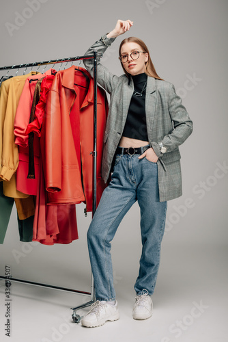 Φωτογραφία attractive stylist in glasses standing with hand in pocket near trendy clothing
