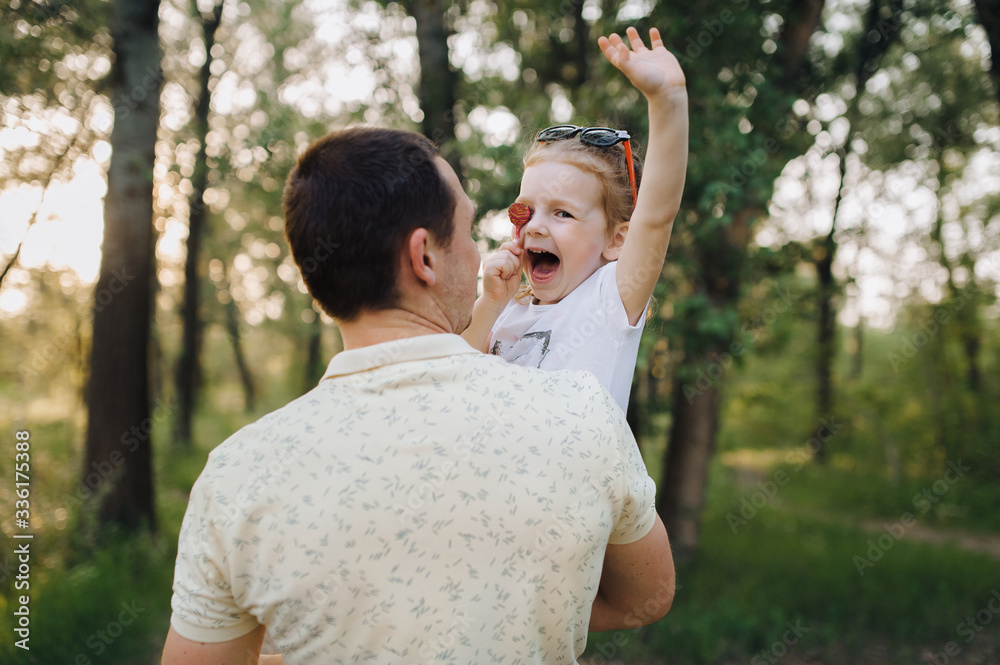 A young man dad in a tracksuit carries in his arms his beloved daughter in nature, who closes her eyes with candy and laughs. Funny and funny photo, concept. A young man dad in a tracksuit carries in his arms his beloved daughter in nature, who closes her eyes with candy and laughs. Funny and funny photo, concept.