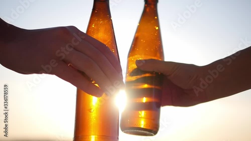 A group of friends clink glasses of beer bottles against the sky and sunset, the bottom corner of the camera. The soft rays of the sun shine through the glass of a bottle