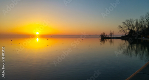 Beautiful sunset over a lake, with flying birds and calm, smooth water, and the pier and some trees.   © Mate