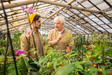 Senior nursery grower holding potted plant and explaining young man how to grow flowers in greenhouse