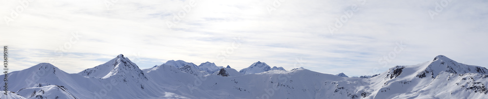 Panorama of high winter mountains with snowy slopes and sunlit cloudy sky