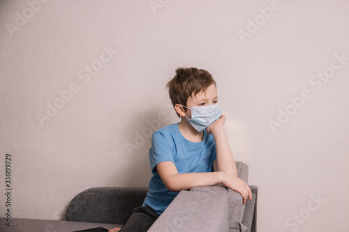 A boy in a medical mask sits on a sofa and is sad