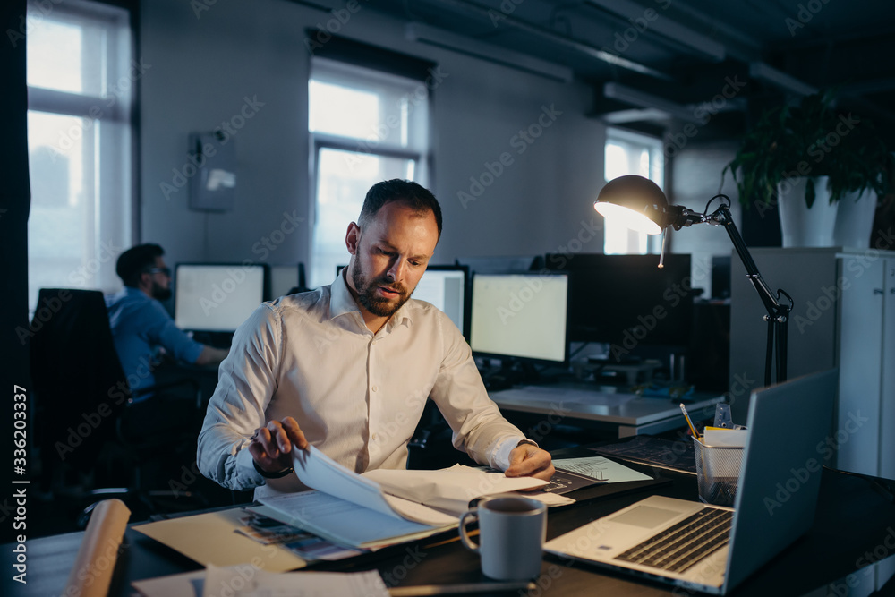 Handsome man in white shirt works with blueprints and documents, sitting at the desk Handsome man in white shirt works with blueprints and documents, sitting at the desk