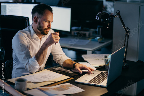 Mature male careerist working at night in the dark office. Light from lamp and monitors