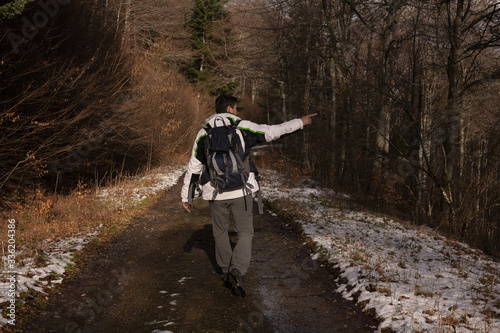 A man with a bagpack is hicking in Giromagny winter forest, and he is showing something 
