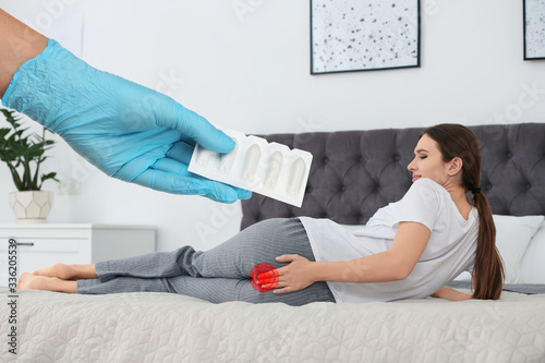 Doctor holding suppositories for hemorrhoid treatment and woman suffering from pain on bed at home