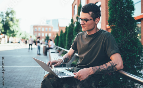 Concentrated male student in spectacles for provide eyes from computer light working with information for university course work connected to public internet in campus, man reading news on website