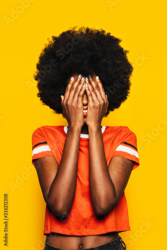 Beautiful Afro Woman posing over yellow background covering her face with hands photo