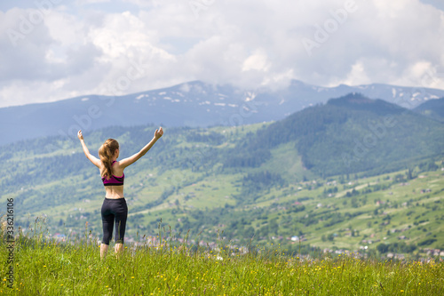 Slim young woman with raised arms outdoors on background of beautiful mountain landscape on sunny summer day.