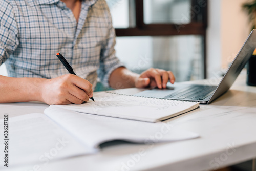 Young Asian man using laptop at home while writing on notebook