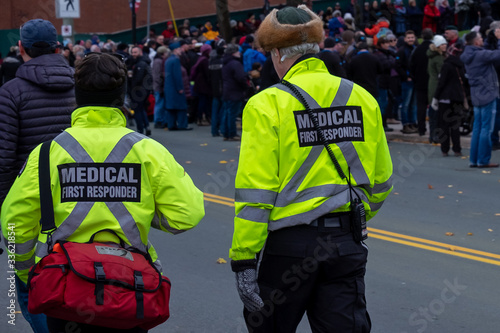 A male and female medical first responder walking on a road among a crowd of people. Both are wearing bright reflective yellow and grey paramedic uniforms with a soft cloth red medical first aid bag.