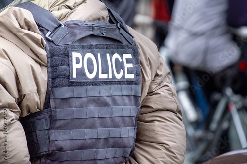 A police officer wearing a khaki parka down-filled uniform and a black bulletproof vest with the word police on the back. The person has their hands behind their back standing at ease.