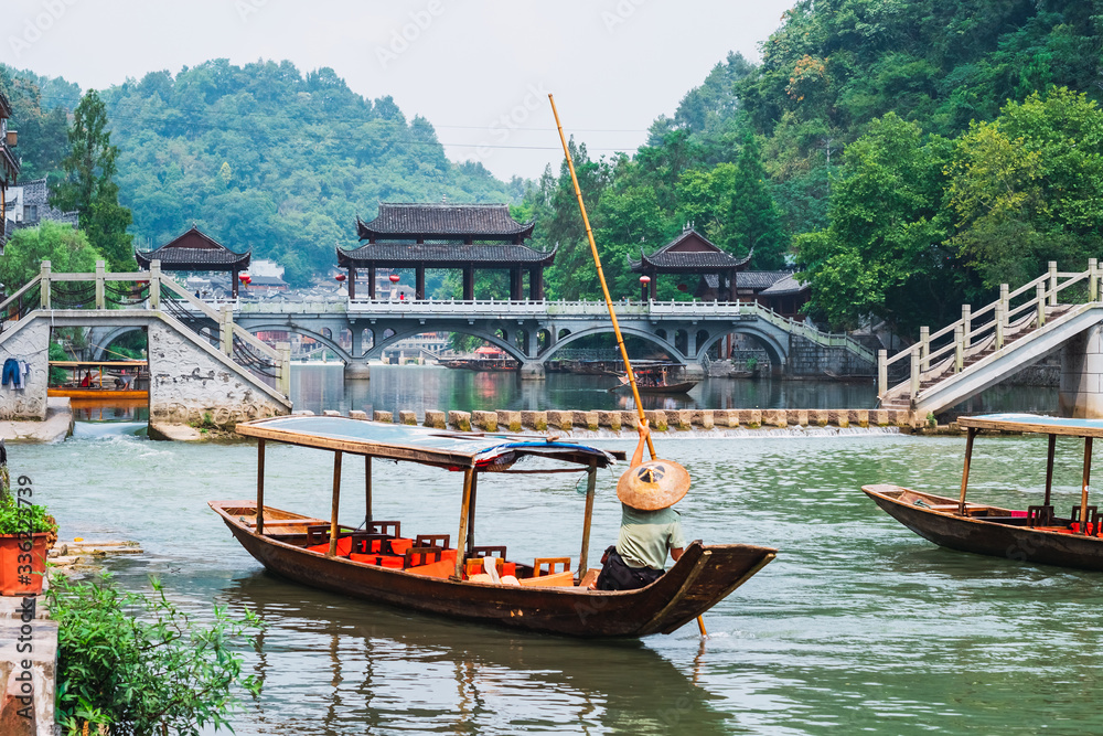 Man in a boat with a traditional triangular chinese hat standing in waters of Tuo river flowing through the centre of ancient city Fenghuang Old Town