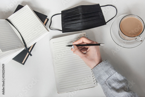 Hand holds a pencil over a notebook. Nearby is a cup of coffee and a black medical mask. Work from home. Stay home in the coronavirus. Pandemic concept. photo