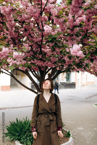 Happy travel woman with backpack, pink sunglass and jeans khaki coat. Sakura cherry blossoms tree on background. Vacation concept. Vertical picture.
