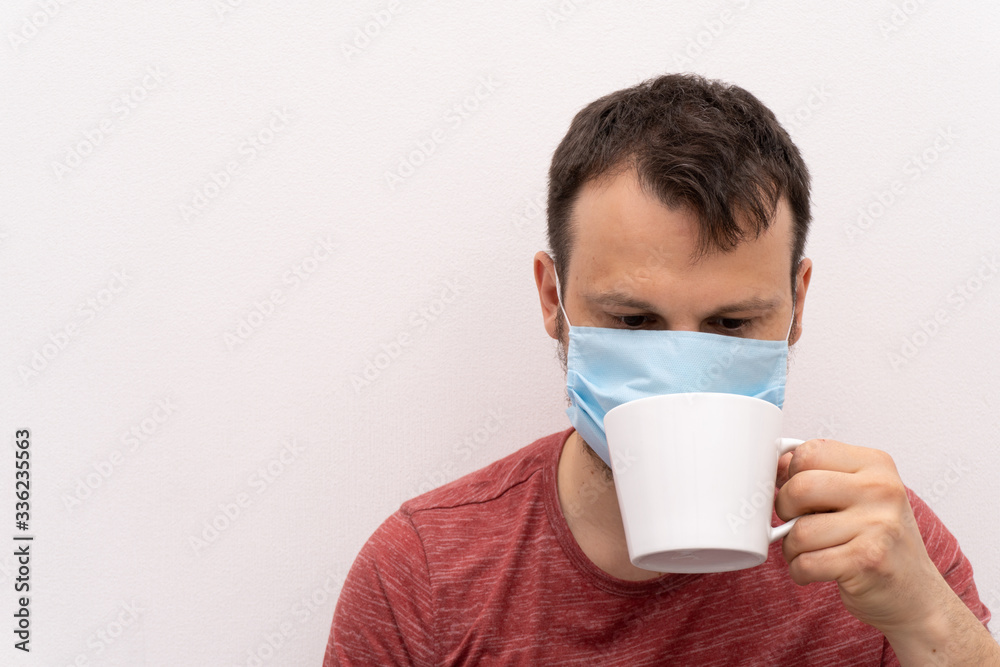 Caucasian man in facial surgical medical protective mask tries to drink coffee or tea from a white cup, white background. Caucasian man in facial surgical medical protective mask tries to drink coffee or tea from a white cup, white background.