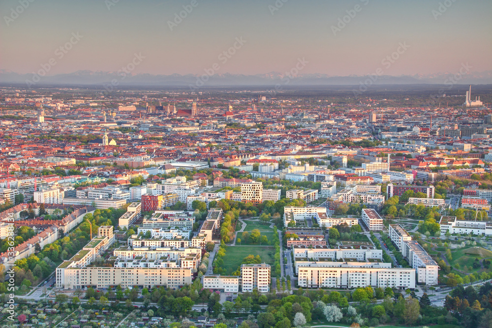 Aerial view of Munich outskirts and historic center Altstadt at sunset with  housing estates, office buildings, green parks, church towers and snowy Bavarian Alps in background, Bayern Germany Europe