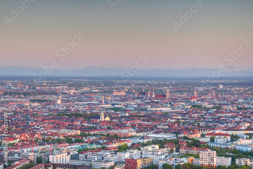 Aerial view of city outskirts and historic center in evening sunlight with church towers, modern residential and commercial buildings and Bavarian Alps in background, Munchen Bayern Germany Europe