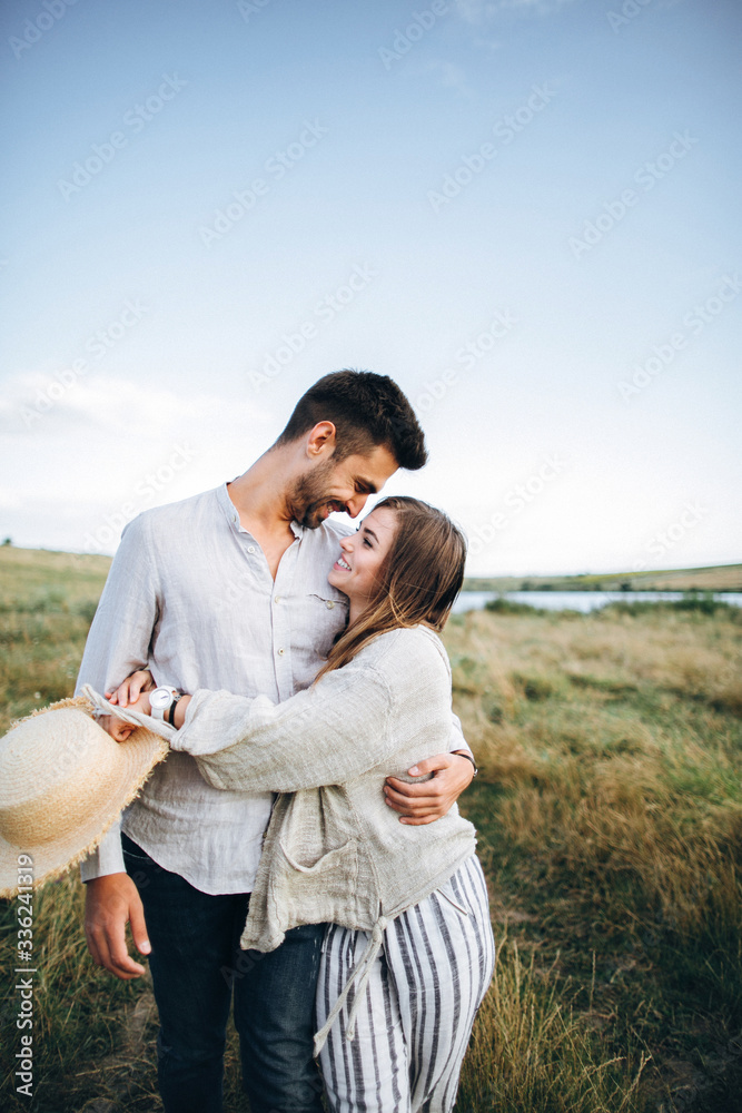 Happy couple in love hugging, kissing and smiling against the sky in field. Hat in girl's hand Happy couple in love hugging, kissing and smiling against the sky in field. Hat in girl's hand