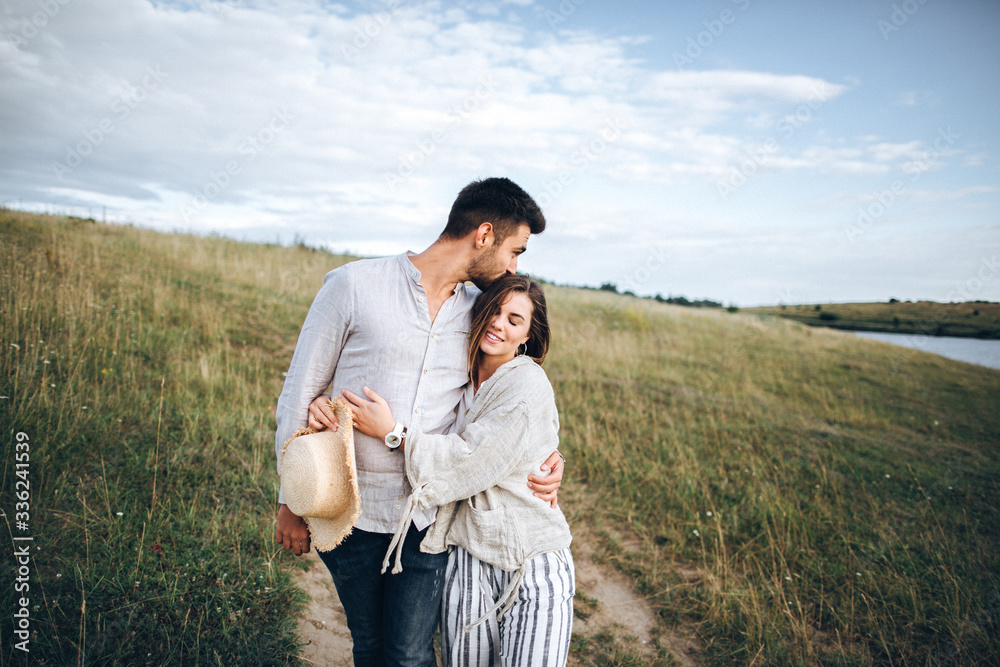 Fototapeta premium Happy couple in love hugging, kissing and smiling against the sky in field. Hat in girl's hand