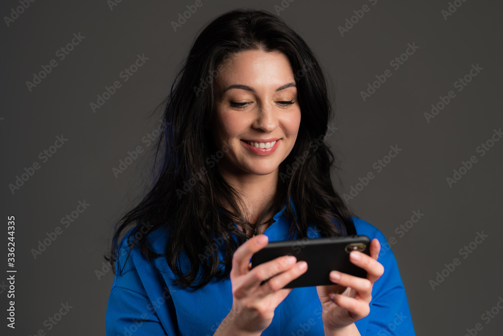 Mature woman sms texting, using app on smartphone. Pretty adult lady surfing internet with mobile phone. Grey studio portrait. Mature woman sms texting, using app on smartphone. Pretty adult lady surfing internet with mobile phone. Grey studio portrait.