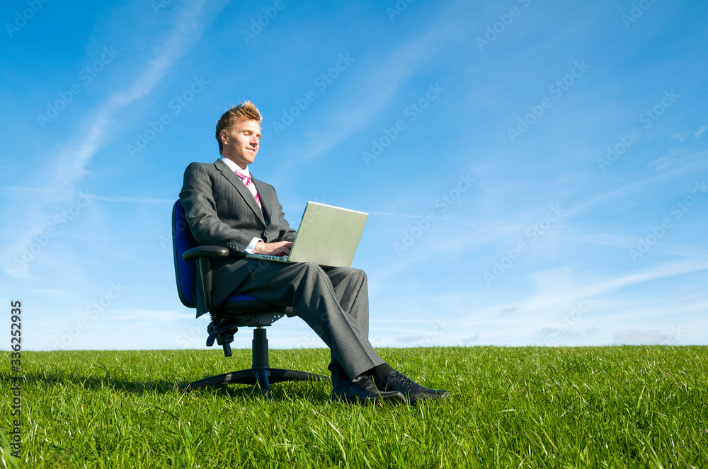 Smiling businessman sitting outdoors on an office chair in a sunny green meadow working on his laptop Smiling businessman sitting outdoors on an office chair in a sunny green meadow working on his laptop