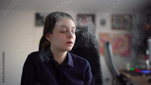 A young girl in a shirt elegantly smokes an electronic cigarette. Exhales white smoke