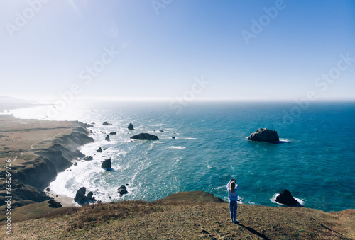 Female traveler making photo of beautiful marine nature environment during wanderlust getaway trip on vacation, back view of hipstergirl explorer standing on rock near ocean water taking photo photo