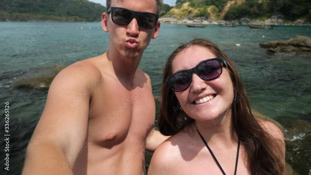 happy young couple in sunglasses makes selfie against beautiful ocean hilly beach at tropical resort close view