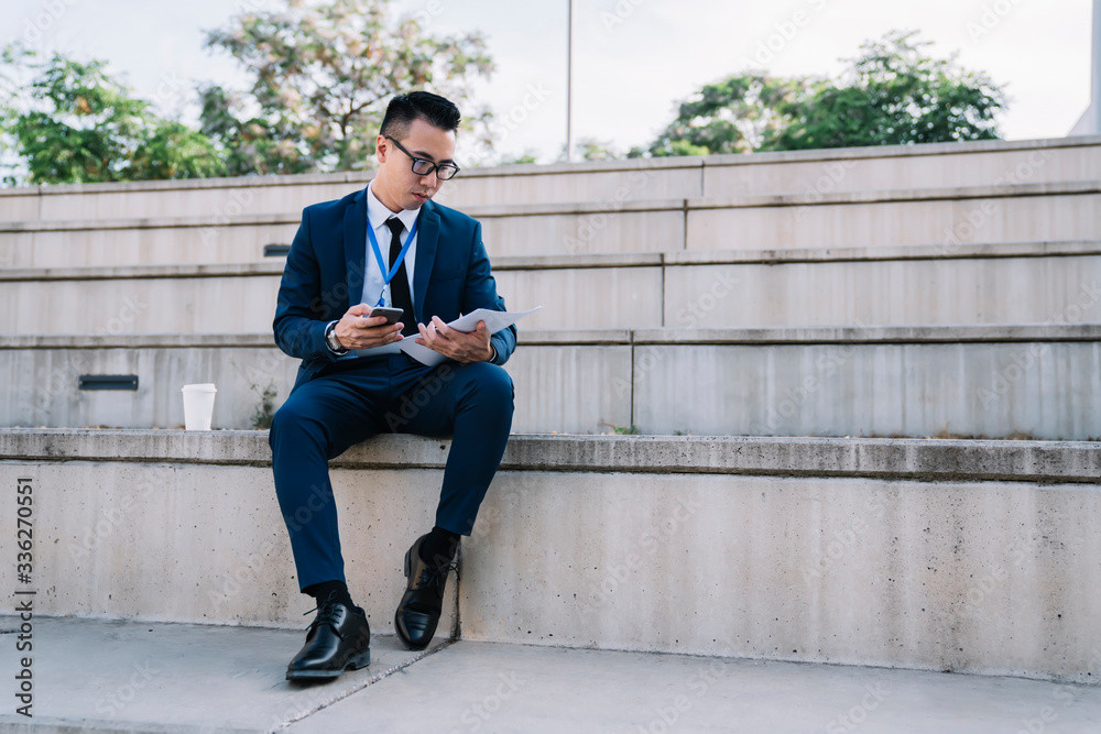 Serious male employee in suit browsing smartphone while analyzing document