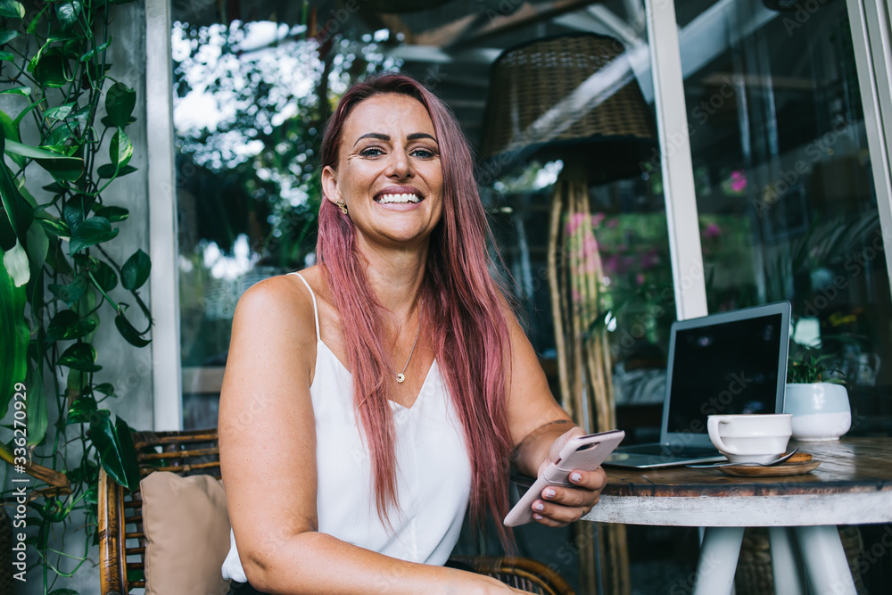 Long haired woman smiling widely holding mobile phone at table in cafe