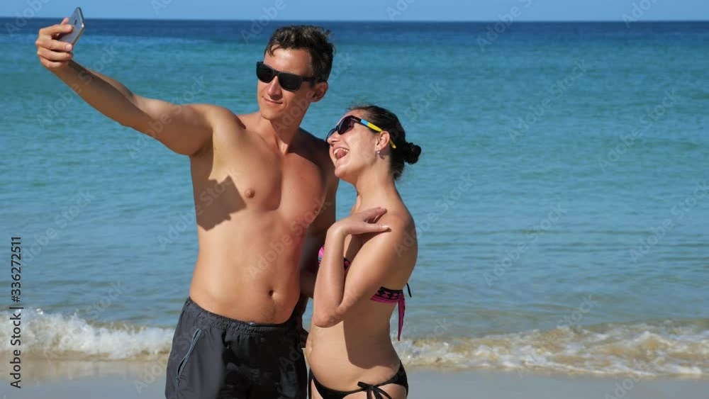 smiling happy couple in swimsuits and sunglasses makes selfie with smartphone on sunny beach smiling happy couple in swimsuits and sunglasses makes selfie with smartphone on sunny beach
