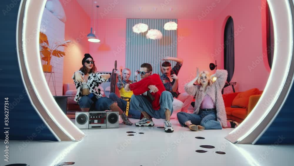 Zoom in shot of young eccentric male and female dancers in hipster outfits sitting on the floor in trendy studio with pink light and dancing vogue to music on vintage cassette tape player