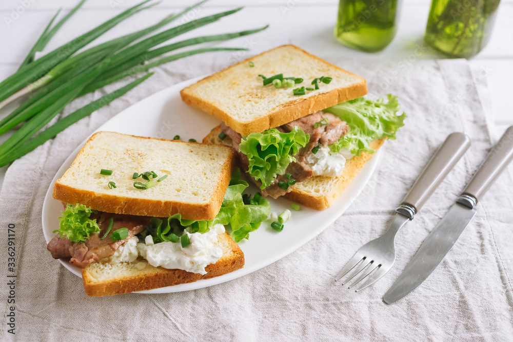 White wheat bread toasted sandwich with pork meat and cheese, salad and sour cream. Chopped chives, bottled olive oil, a knife and a fork. Against the background of a gray fabric napkin.