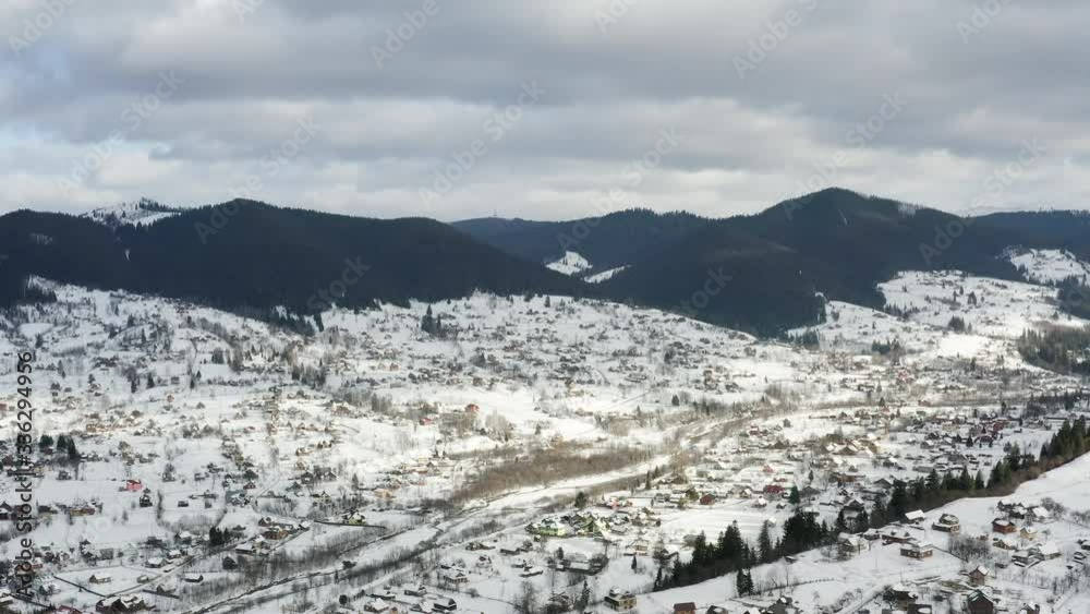 Aerial view of a Mountain Village with Hills Covered in Snow and Pine Forest in Winter, Ukraine. Flying over the snowy hills and wooden small houses. Countryside, fir tree. Drone view.