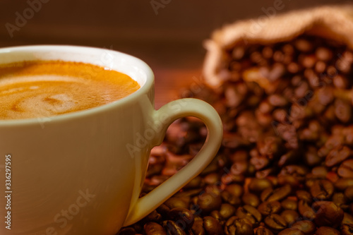 Close-up shot of hot fresh coffee or cappuccino for the morning with milk foam in a white ceramic cup with dark brown coffee beans roasted fragrance in a sack on table background with copy space.