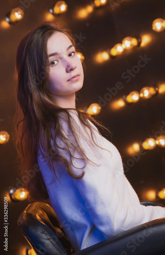 A beautiful young girl is sitting on a chair against the background of a wall with light bulbs