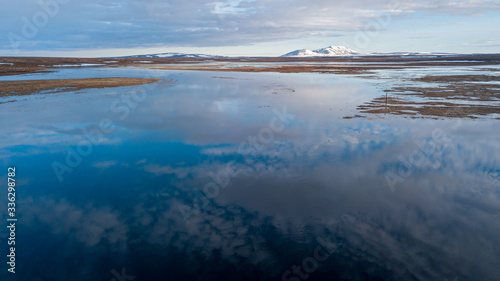 calm water reflects blue sky with white clouds against brown meadows and snowy hillpeak on horizon