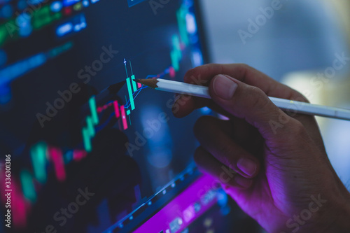 Business man hand holding pencil points to stock market graph analysis on monitor screen. Economic and financial concept. Seletive focus.