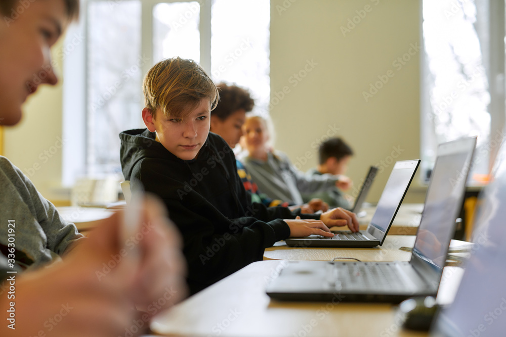Seeking help. Portrait of caucasian schoolboy looking at the laptop of his neighbor while sitting in a classroom with other pupils during a lesson in modern smart school Seeking help. Portrait of caucasian schoolboy looking at the laptop of his neighbor while sitting in a classroom with other pupils during a lesson in modern smart school