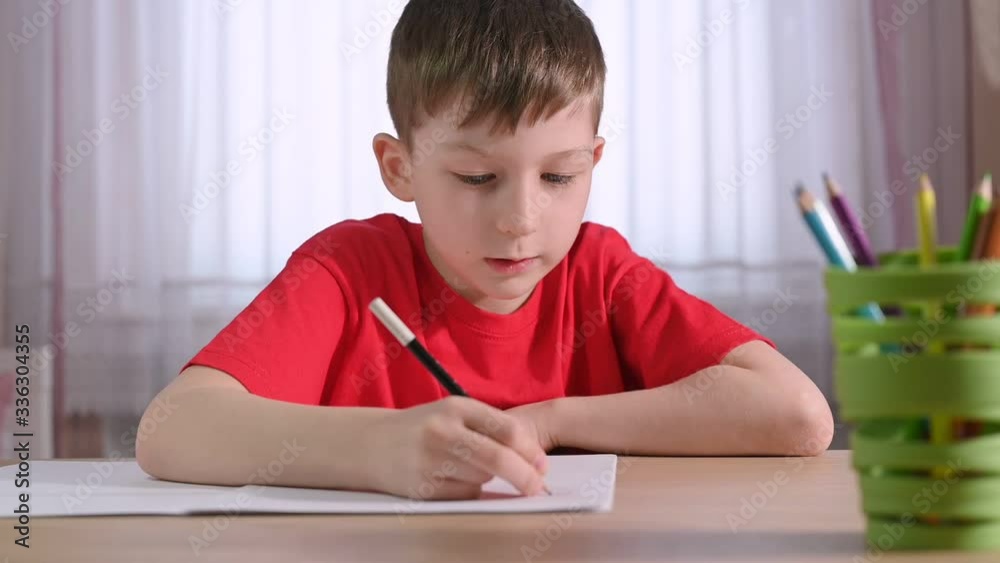 serious boy in red t-shirt writes homework in exercise book at table with green pen holder against bright curtain closeup serious boy in red t-shirt writes homework in exercise book at table with green pen holder against bright curtain closeup