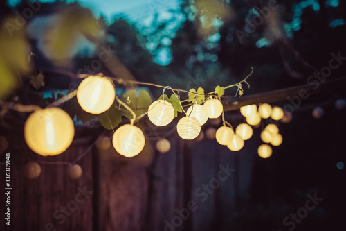 Row of round paper lanterns in a home garden on a wooden thatched background at night. Festive decoration in the garden on an evening.