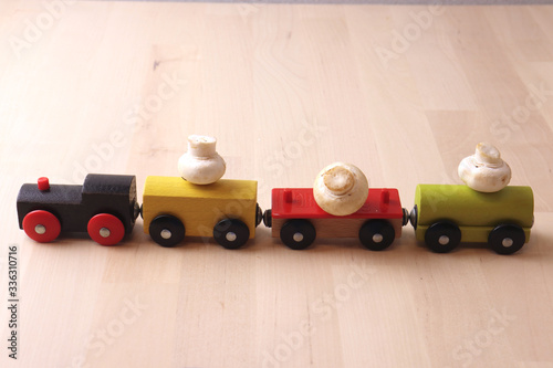  toy, wooden, multi-colored train transports a crop of champignon mushrooms, on a light wooden background. Delivery of products in quarantine, vitamins, immunity, diet, proteins