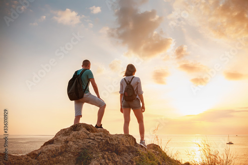Two hikers stands on big rock and looks at sunset sea