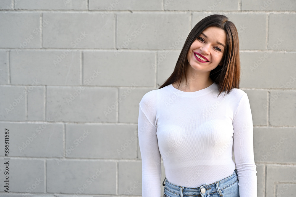 A caucasian portrait of a pretty brunette girl in a white blouse stands near the wall of a building on a spring day in the city. Model with pleasure and smile posing.
