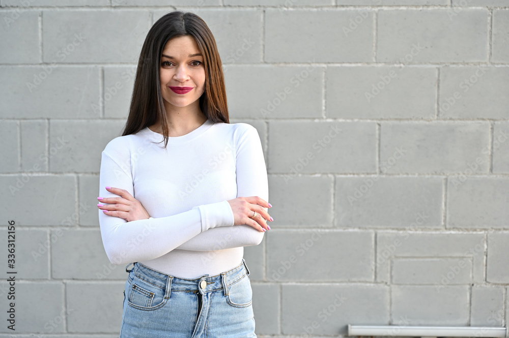 A caucasian portrait of a pretty brunette girl in a white blouse stands near the wall of a building on a spring day in the city. Model with pleasure and smile posing. A caucasian portrait of a pretty brunette girl in a white blouse stands near the wall of a building on a spring day in the city. Model with pleasure and smile posing.